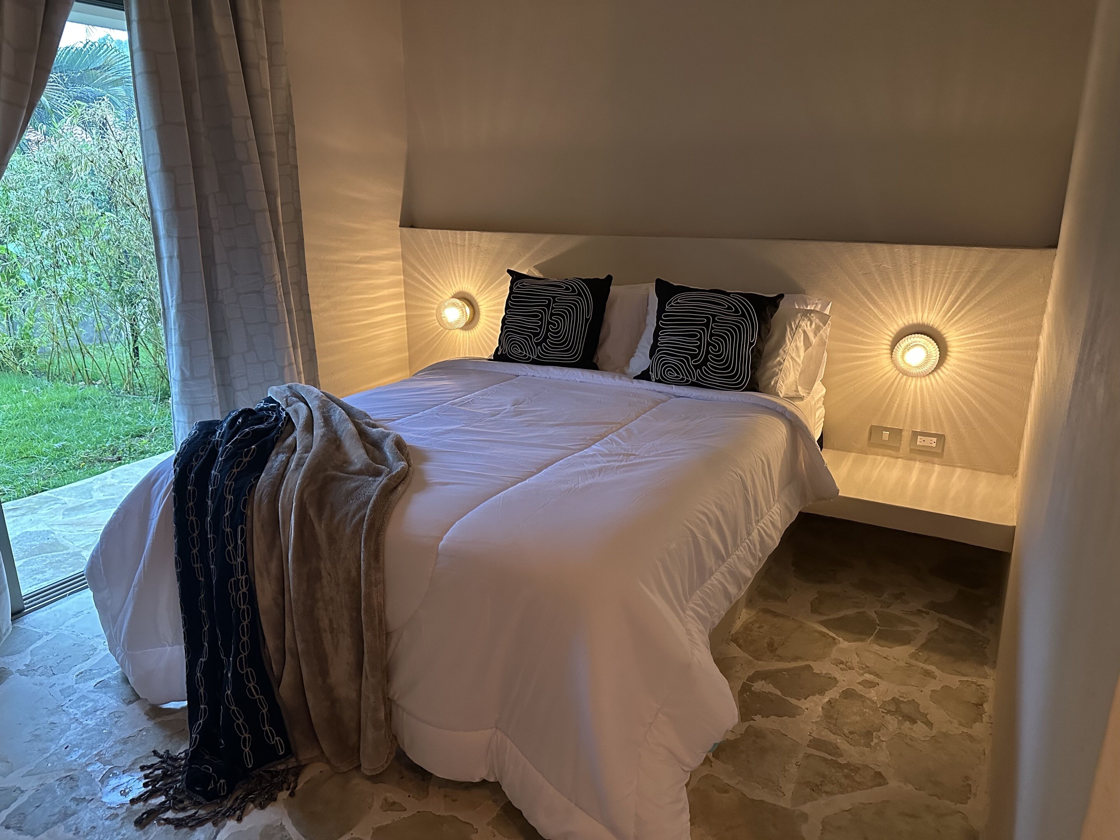 Ett sovrum med en stor säng, två sänglampor, utsikt över grönområden utanför och ett stengolv.