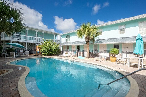 A swimming pool with lounge chairs and umbrellas, surrounded by a building with a light blue exterior and a covered patio area.