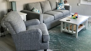 Olohuoneessa on harmaa sohva, harmaa nojatuoli, valkoinen sohvapöytä ja kaksi seinälle asennettua lamppua. Seinällä on kaksi taulua, katossa kattotuuletin ja ikkunassa sälekaihtimet.
