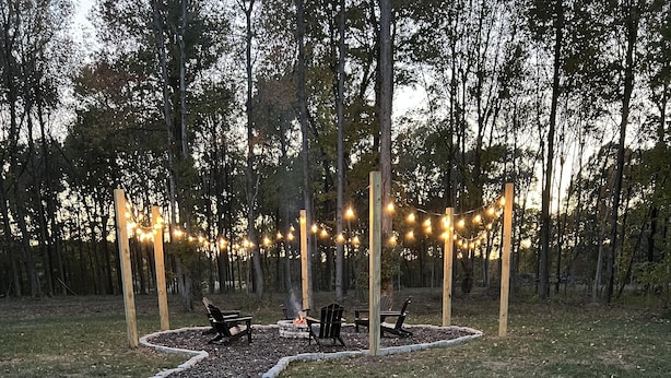 Un sendero de grava bordeado por piedras que conduce a una zona de estar con bancos y un hogar para la leña, rodeada de árboles y luces colgantes.