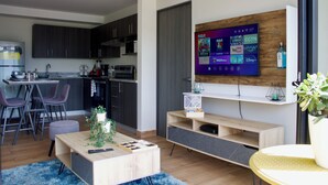Una sala de estar moderna con un televisor de pantalla plana montado en una pared de madera, un comedor con una cocina al fondo y una pequeña zona de estar con una mesa de centro y sillas.