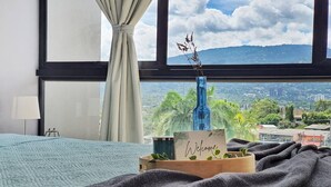 Habitación de hotel con un ventanal grande, una cama con colcha azul, una manta gris oscuro, un letrero de bienvenida de madera y un jarrón azul con una planta.