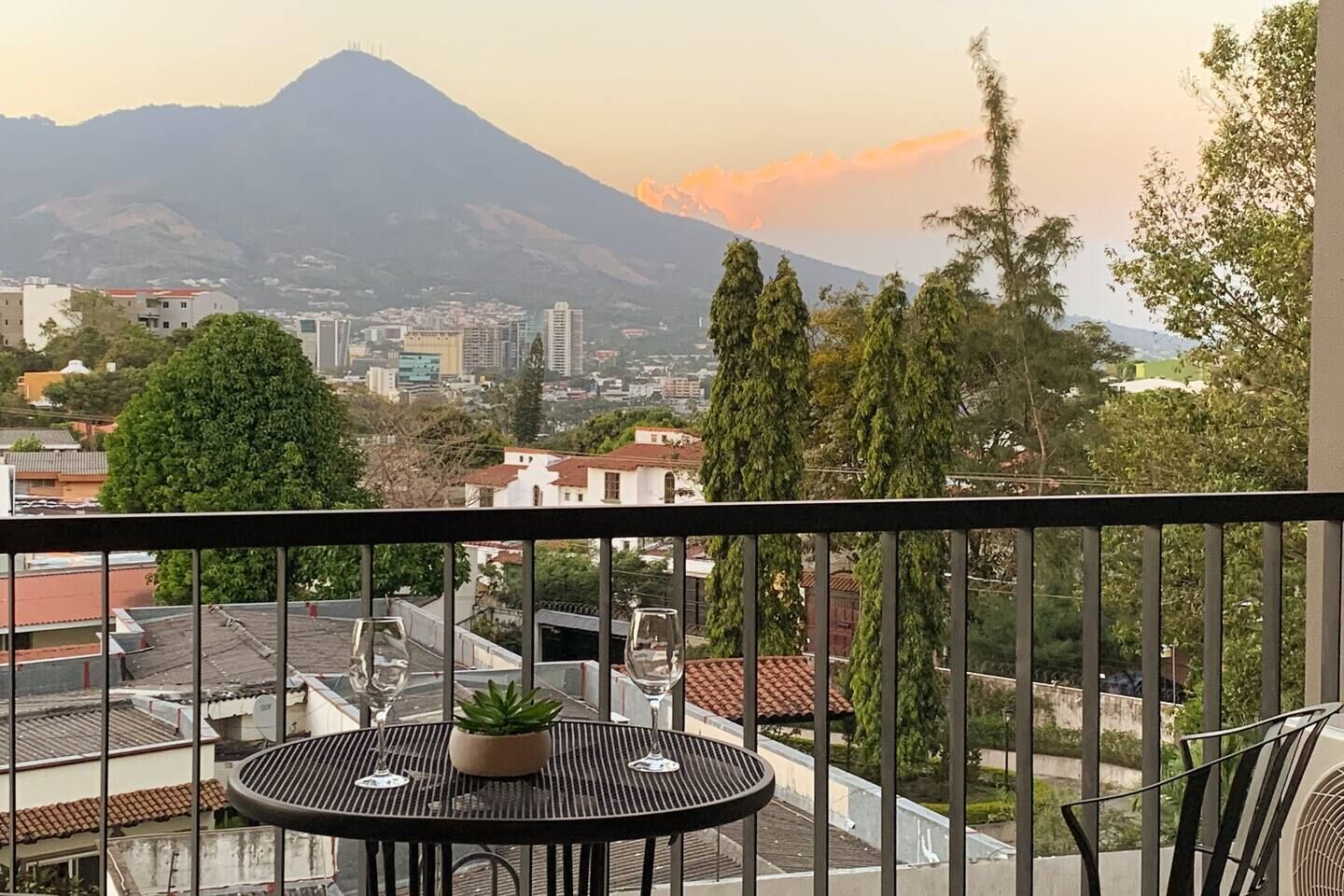 A balcony with a table and chairs, overlooking a cityscape with buildings and a mountain in the background.
