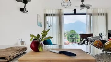 Een keuken met een houten snijplank, een mes en een potplant. Er is een eethoek met een tafel en stoelen, en een uitzicht op bergen door het raam.