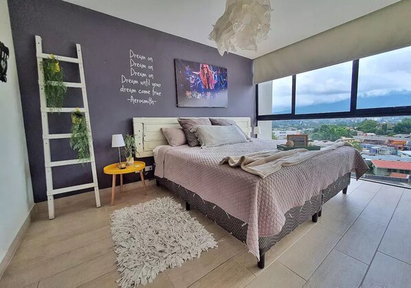 Camera moderna con un letto grande, vista sulla città, una scala con piante verdi e una citazione motivazionale sulla parete.