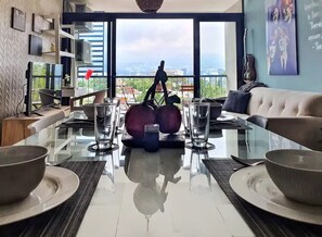 Zona pranzo con tavolo in vetro, piatti, bicchieri, un elemento decorativo rosso e vista sulla città attraverso ampie finestre.