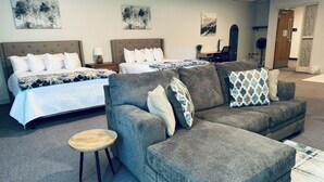 Une chambre d’hôtel avec un grand lit, un canapé gris et un petit tabouret en bois.