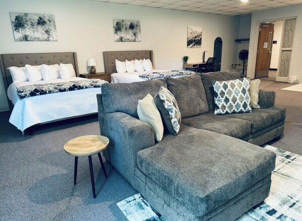 A hotel room with a large bed, a grey sofa, and a small wooden stool.