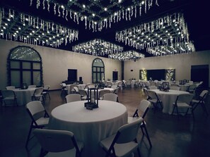 A spacious banquet hall with round tables and chairs, decorated with hanging lights and a chandelier.