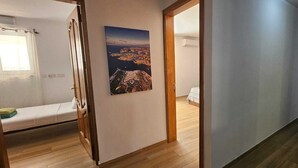 Un couloir avec un plancher en bois, un lit avec un matelas blanc, une fenêtre avec un rideau à motifs et un tableau représentant un paysage côtier.