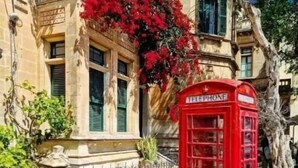 Une cabine téléphonique rouge se trouve devant un immeuble à la façade en pierre et aux volets verts.