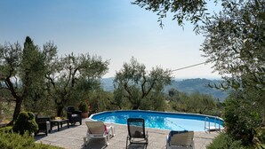 Ein Swimmingpool mit Liegestühlen und einem Tisch, umgeben von Bäumen und Blick auf ferne Hügel.