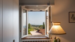 Ein Zimmer mit Fenster und Blick auf eine Landschaft, ein Heizkörper unter dem Fenster sowie eine Lampe mit grünem Fuß auf einem Tisch.