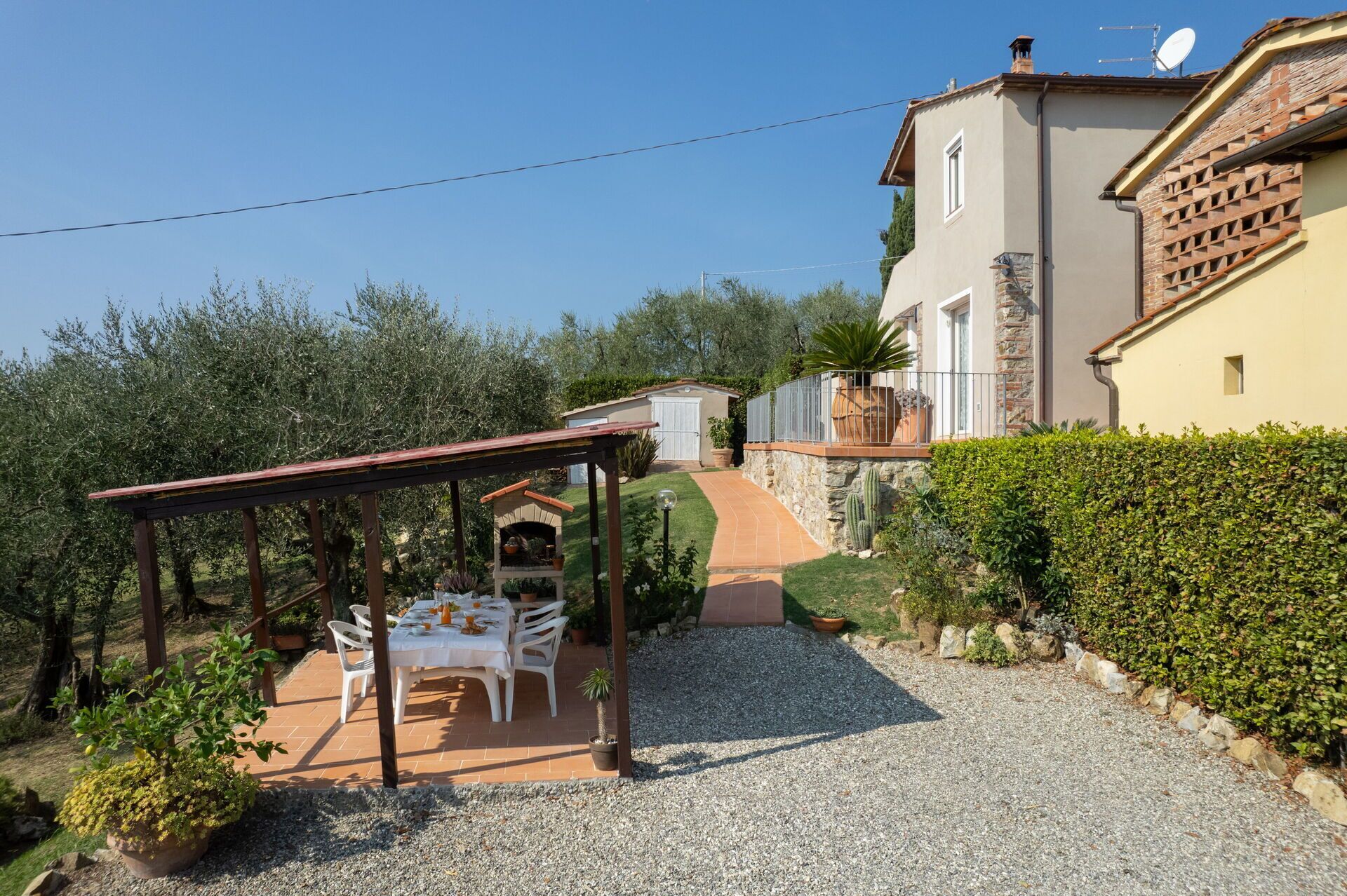 Une terrasse avec une table et des chaises, un espace salon abrité et un chemin de gravier menant à une maison dotée d’un mur en pierre et d’une antenne parabolique.