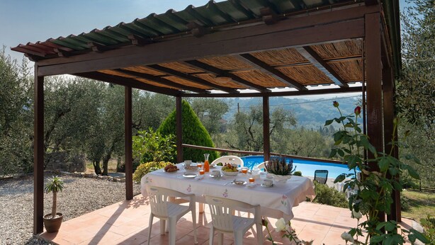 Ein überdachter Essbereich im Freien mit einem für das Frühstück gedeckten Tisch, mit Blick auf einen Pool und eine Berglandschaft.