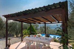 Un área de comedor al aire libre techada, con una mesa lista para el desayuno, con vista a una piscina y un paisaje montañoso.