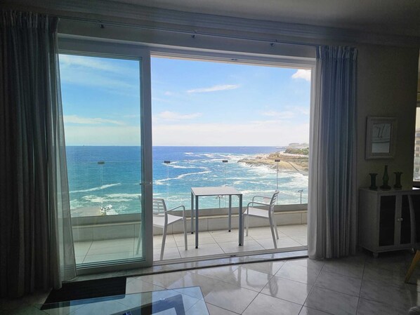 A room with a large sliding glass door leading to a balcony with a view of the ocean and rocky coastline. - Seaside Serenity 3BR Oasis Breathtaking Views (Tas-Sliema)