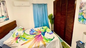 Une chambre à coucher comprenant un lit, un ventilateur de plafond, une fenêtre avec des rideaux, un climatiseur fixé au mur et une porte avec un volet en bois.