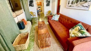 Un salon avec un canapé rouge, une table basse en bois et un tapis à motifs verts. On y trouve deux fauteuils, une petite table et un téléviseur fixé au mur.