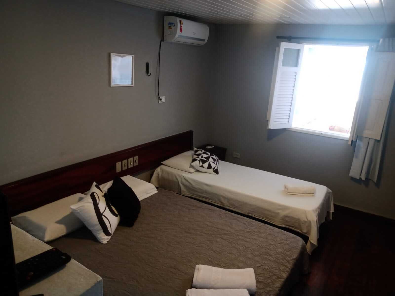 A hotel room with two beds, a window, an air conditioning unit, and a bedside table with a remote control.
