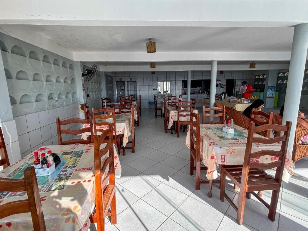 Un restaurante con sillas y mesas de madera, suelo de baldosas y una zona de cocina al fondo.