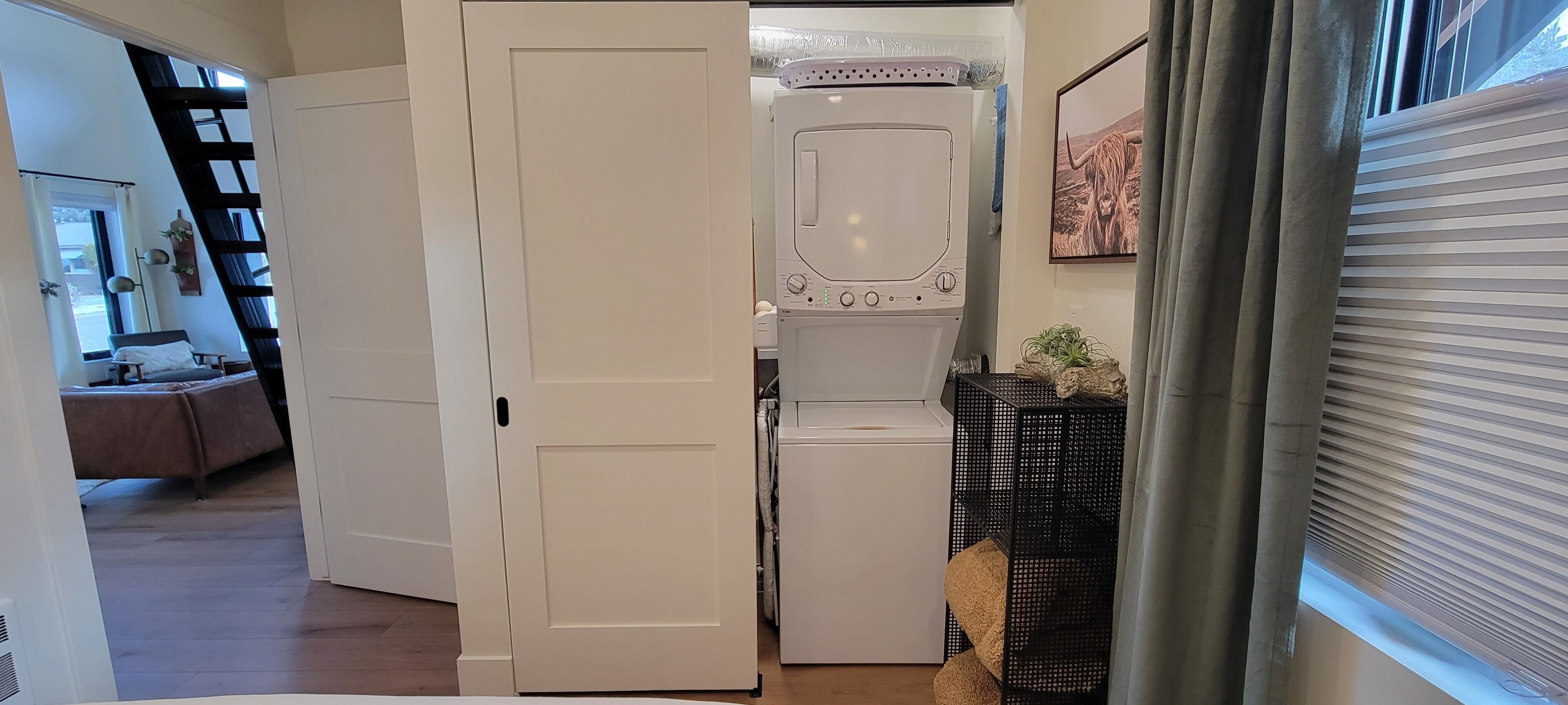 Une buanderie équipée d’un lave-linge et d’un sèche-linge, une porte donnant sur une chambre, et une fenêtre avec des stores.
