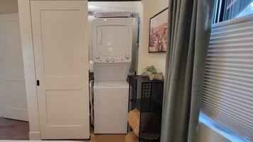 Une buanderie équipée d’un lave-linge et d’un sèche-linge, une porte donnant sur une chambre, et une fenêtre avec des stores.