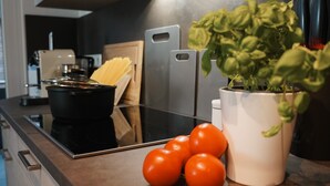 A kitchen counter with a black induction cooktop, a white pot, a potted basil plant, and a bunch of tomatoes. - Paderborn Oasis with Fireplace, Parking & Balcony, Stylish Home, 24/7 Check-in (Paderborn)