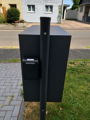 Une boîte aux lettres noire, placée à l’extérieur, avec un trou pour la clé, située sur une surface pavée, avec de l’herbe et un immeuble d’habitation en arrière-plan.