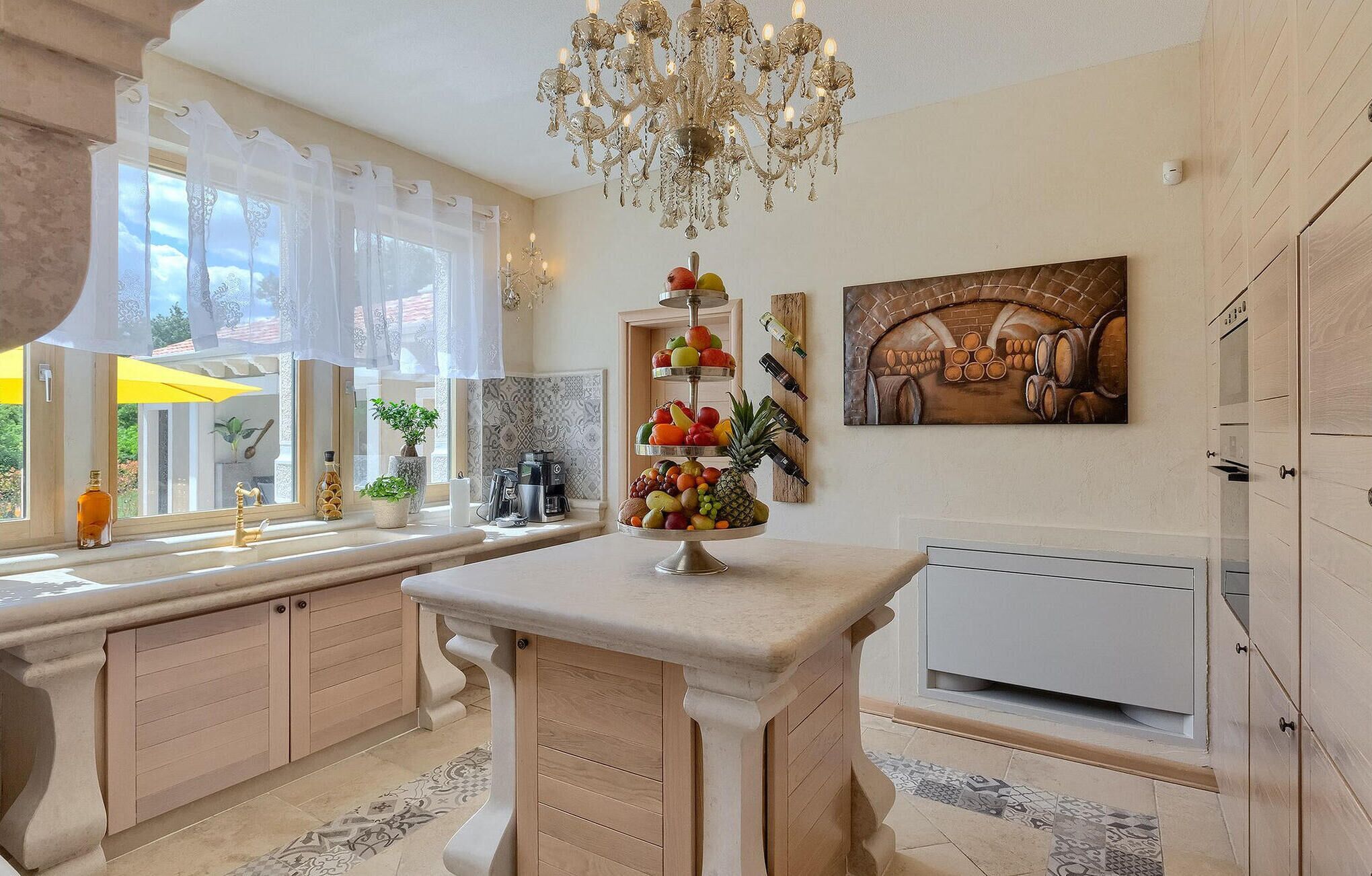 A kitchen with a central island, a chandelier, a fruit display, and a wine rack.