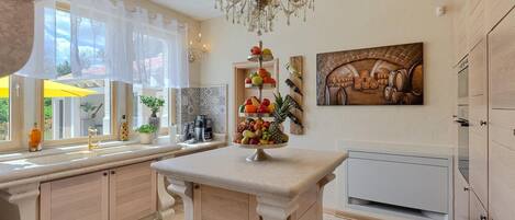 A kitchen with a central island, a chandelier, a fruit display, and a wine rack.