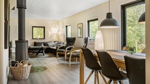 Ein modernes Wohnzimmer mit einem großen schwarzen Herd, Holzböden, einer Essecke mit Tisch und Stühlen sowie großen Fenstern mit Blick auf Grünflächen.