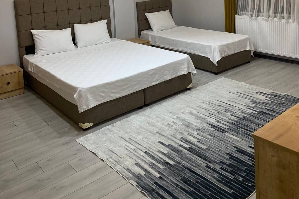 A modern hotel room with two beds, a large window with sheer curtains, a wooden floor, and recessed ceiling lighting.