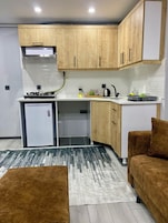 Una cocina compacta con armarios de madera, horno empotrado y un refrigerador pequeño. En la sala de estar, hay un tapete gris y dos sofás marrones.
