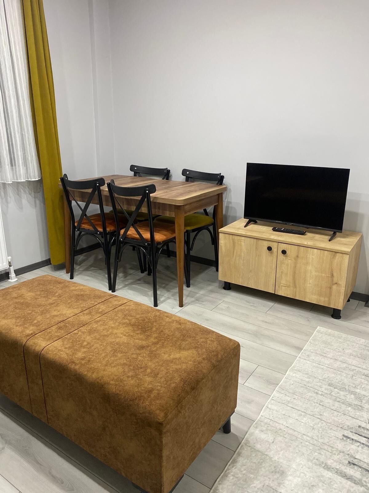 Una sala de estar con una mesa de comedor de madera, dos sillas, un sofá, un televisor sobre un mueble de madera y una cortina amarilla.