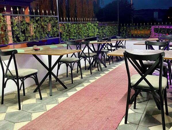An outdoor seating area with multiple tables and chairs, a red carpet, and decorative plants along the walls.