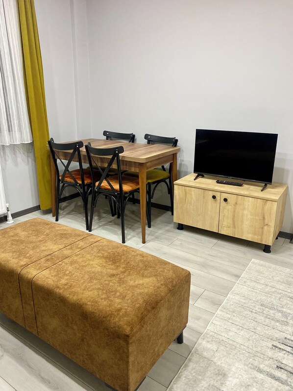 Una sala de estar con una mesa de comedor de madera, dos sillas, un sofá, un televisor sobre un mueble de madera y una alfombra de color claro.