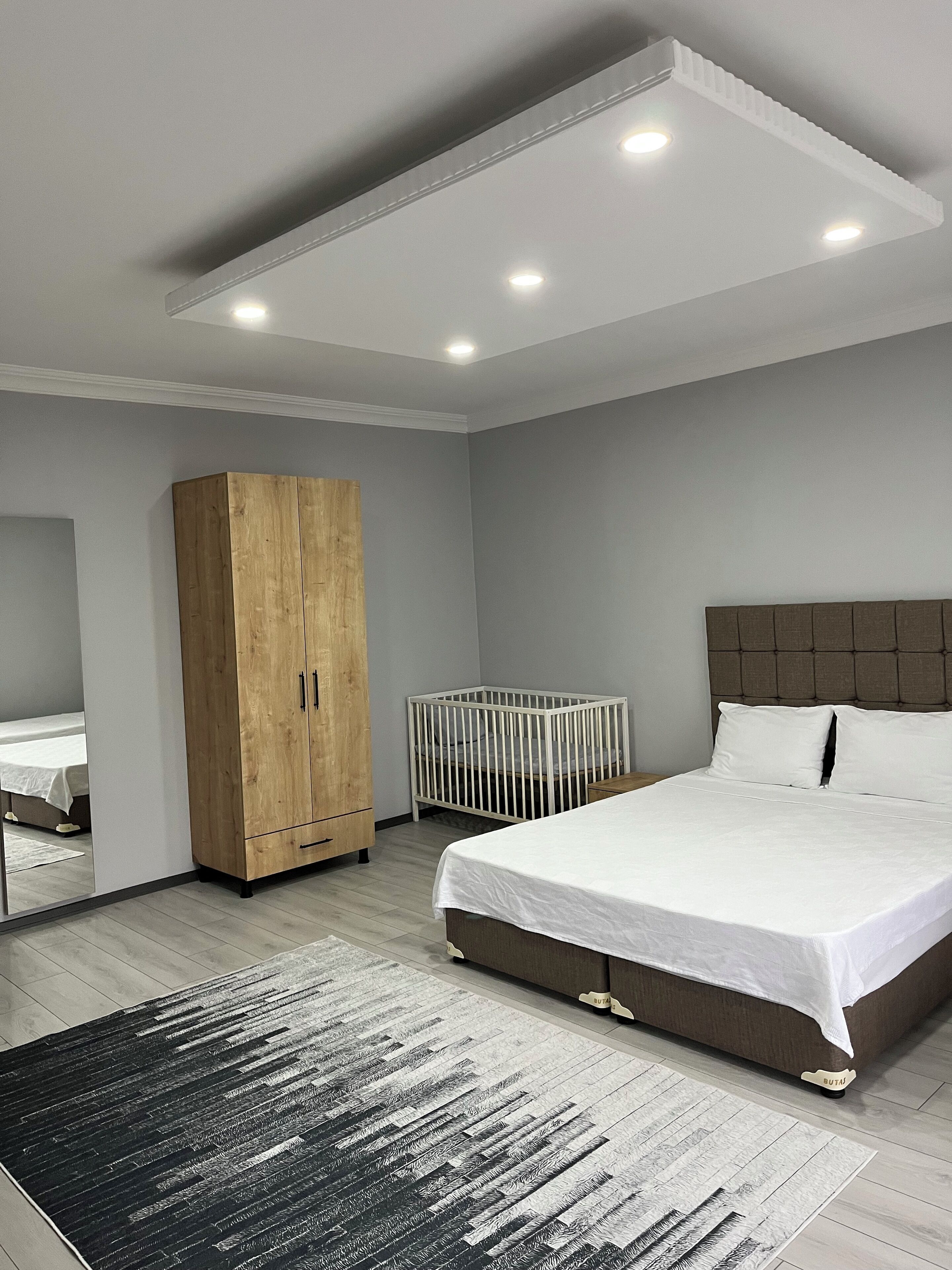 Un dormitorio moderno con una cama grande, un armario de madera, una cuna y un espejo.