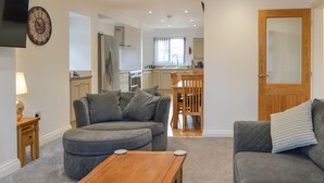 Ein modernes Wohnzimmer mit einem grauen Ecksofa, einem Holztisch und einem grauen Sessel. Im Hintergrund ist eine Küche mit weißen Schränken und einem Fenster erkennbar.