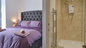Ein modernes Schlafzimmer mit einem gesteppten Kopfteil, einem großen Bett mit violetter Bettwäsche und einem Duschbereich mit Glastrennwand sowie einer Handbrause.