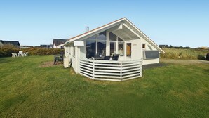 A white, single-story house with a large glass window, a deck, and a small outdoor seating area. - Nice home in Frøstrup with sauna (Frøstrup)