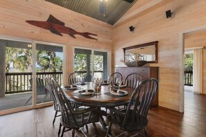 Comedor de madera con una mesa redonda, sillas negras y un ventanal con vistas a una terraza con palmeras.