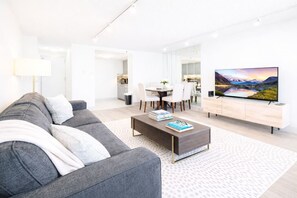 Un salon moderne avec un canapé gris, une table basse sur laquelle sont posés des livres, un téléviseur sur un meuble en bois, et un coin repas à l’arrière-plan.