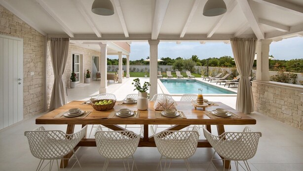 A dining area with a pool view, featuring a wooden table, white chairs, and a stone wall. - Villa Simona Residence (Zminj)