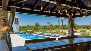 Un área junto a la piscina con mesa de madera, sillas de descanso, pared de piedra y vistas a un edificio moderno y follaje.