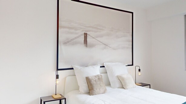 Une chambre à coucher moderne avec un grand lit, du linge de lit blanc et un tableau encadré représentant un pont, accroché au mur.