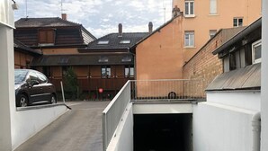 L’entrée d’un parking, avec une rambarde en métal, une voiture garée à l’intérieur et des bâtiments en arrière-plan.
