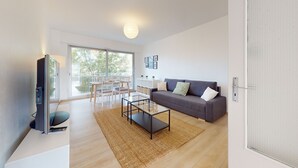 Un salon moderne avec un canapé gris, une table basse en verre et un meuble pour la télévision. Il y a un balcon avec vue sur les arbres.