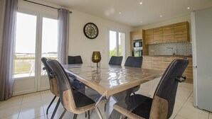 A dining area with a glass table and chairs, a kitchen with wooden cabinets, and a view of the outdoors. - Le Paradis de L'orient (PUITS ET NUISEMENT)