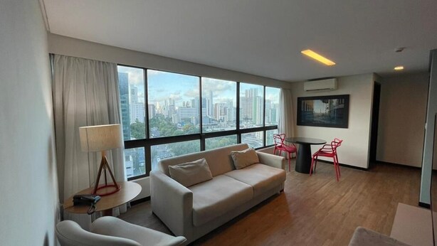 A modern hotel room with a large window offering a city view, a sofa, a small table with a lamp, and a red chair. - Flat Aconchegante em Boa Viagem (Recife)